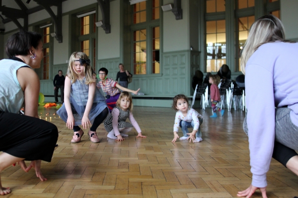 Image shows three young girls mid-dance, crouching on the ground, in a large hall with parquet flooring and green painted walls.