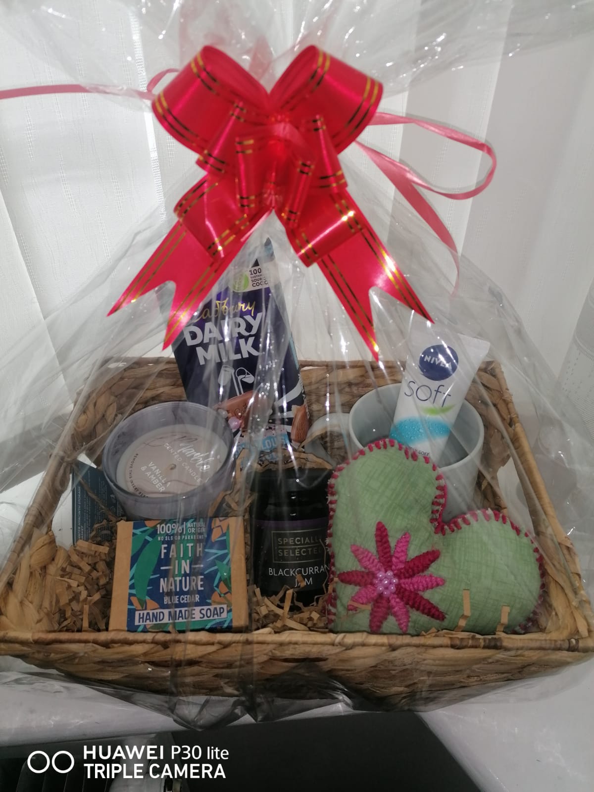 a plastic-wrapped hamper basket containing a candle, chocolate, mug, hand cream, heart-shaped pillow, jam, and soap, with a red ribbon on top