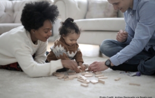 The photo shows a mixed race family - mum, dad, and baby - playing together on the floor.
