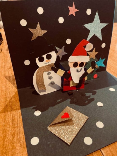 This is a photo of a pop-up card showing Santa and a snowman beside a Christmas tree. The text at the bottom-left reads 