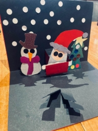 This is a photo of a pop-up card showing Santa and a snowman beside a Christmas tree. The text at the bottom-left reads "Artwork by N Khan".