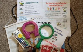 contents of the Explorer Backpack, which includes the newsletter, a notebook, pens, violet magnet, green magnifying glass, plastic insects, modelling clay, and the Explore Manchester Passport