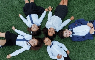 Five smiling children lying down on grass, looking up at the camera