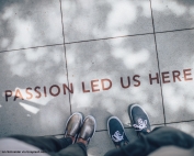 Two people standing on a pavement where the words "PASSION LED US HERE" are written in brown/red paint. The image looks as if the photo was taken by one of the subjects looking down, so that only their legs & shoes, and the pavement, are seen.