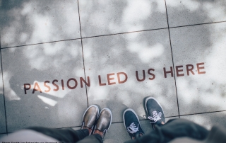 Two people standing on a pavement where the words "PASSION LED US HERE" are written in brown/red paint. The image looks as if the photo was taken by one of the subjects looking down, so that only their legs & shoes, and the pavement, are seen.
