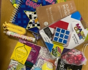 A collection of items that will be going into the parent/carer well-being packs, randomly sprawled on a table. The items include: crossword book, calm colouring book, bath bombs, shea butter masque, colouring pencils, stretchy stress-release toy, heart-shaped chocolates and sweets, hot chocolate sachet, puzzle cube