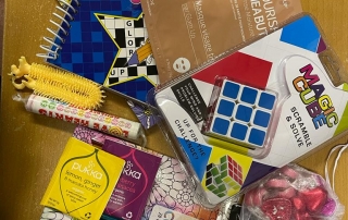 A collection of items that will be going into the parent/carer well-being packs, randomly sprawled on a table. The items include: crossword book, calm colouring book, bath bombs, shea butter masque, colouring pencils, stretchy stress-release toy, heart-shaped chocolates and sweets, hot chocolate sachet, puzzle cube