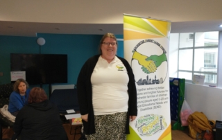 Dawn from our MPCF Team poses beside the MPCF banner, as educational psychologists from One Education speak to parents behind her. This photo was taken at the SEND Local Offer Drop-in in Moss Side Millenium Powerhouse on October 2022.