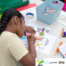 The photo shows a girl colouring a sheet with an illustration of a house. There are other crafts equipment, such as sellotape, markers, glue, and paper on the table. The upper-right side of the photo shows the Z-Arts logo. The lower-right side of the image shows the MPCF, 4CT, 2022 Our Year, and Local Offer logos, respectively.