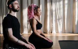 The photo shows a man and a woman sat on the floor, meditating.