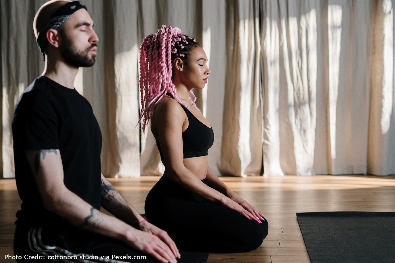 The photo shows a man and a woman sat on the floor, meditating.