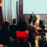 The photo shows a seated man speaking in front of an audience.