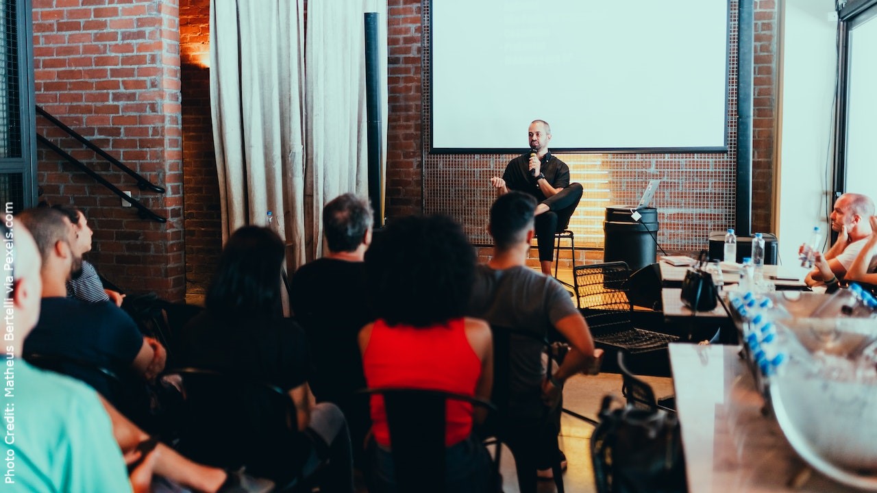 The photo shows a seated man speaking in front of an audience.