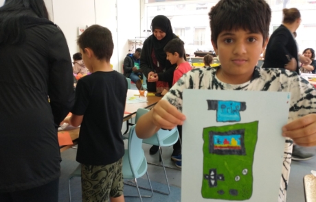 The photo shows a boy proudly showing his artwork while other families enjoy the Relaxed Creative Session at Manchester Art Gallery in summer 2023.