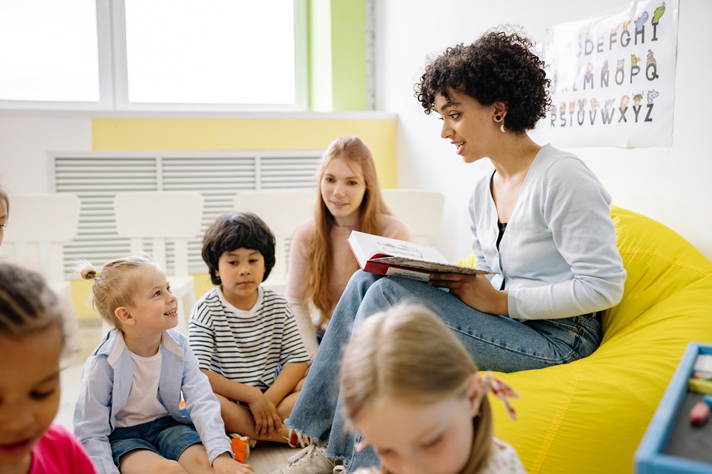 The photo shows a pre-school storytelling session, with a teacher sat reading a book and Early Years children surrounding her.